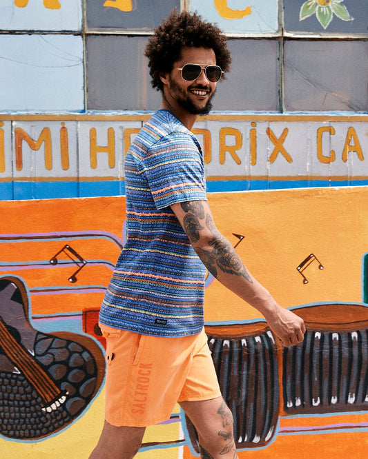 A man with curly hair and sunglasses smiles as he walks past a colorful mural featuring musical instruments and Aztec print, wearing a Saltrock Marks - Mens Short Sleeve T-Shirt in Purple.