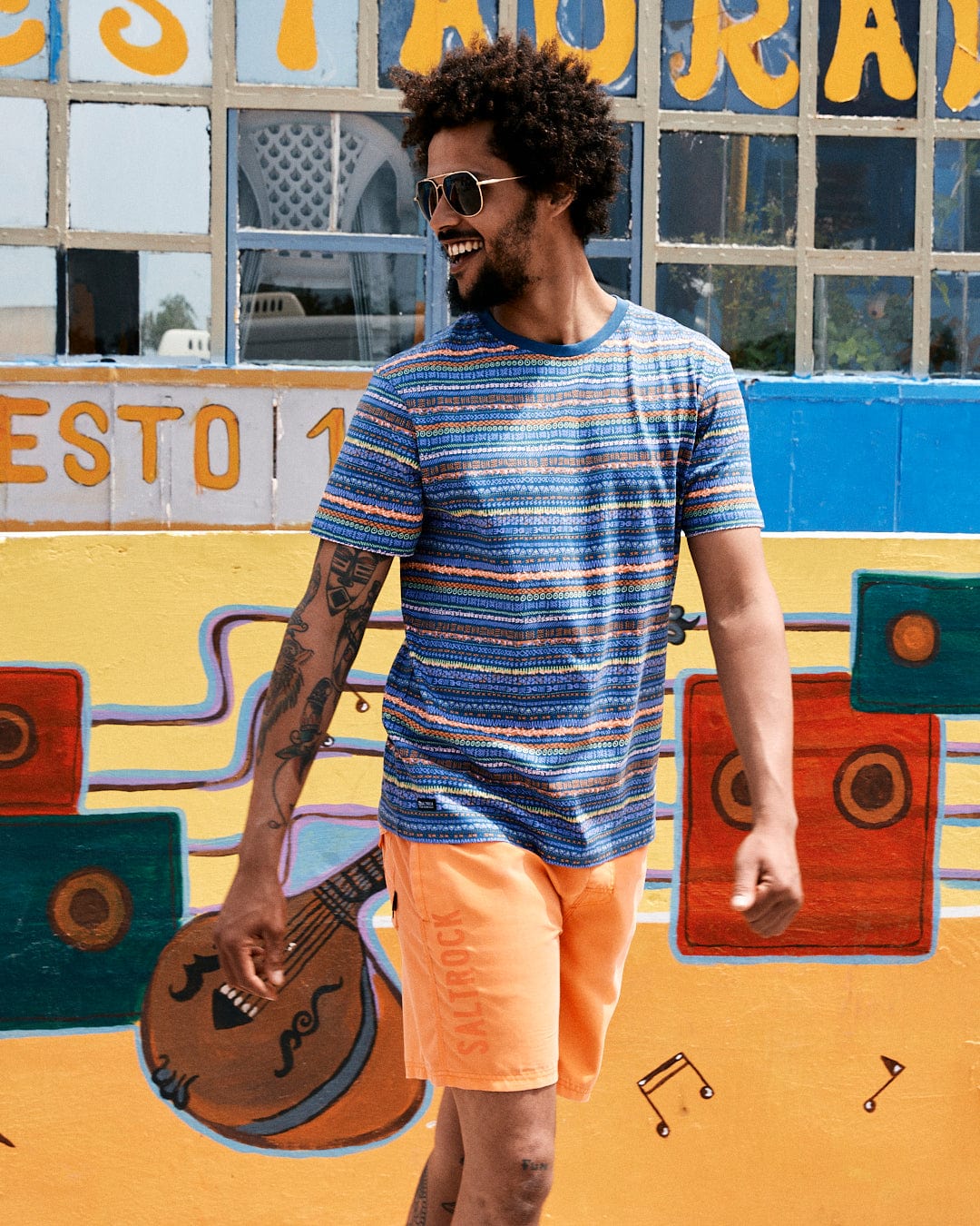 A man with curly hair, wearing sunglasses, smiles as he walks by a colorful mural featuring musical elements. He wears a striped t-shirt with Saltrock branding and orange Marks Mens Short Sleeve T-Shirt - Purple shorts.