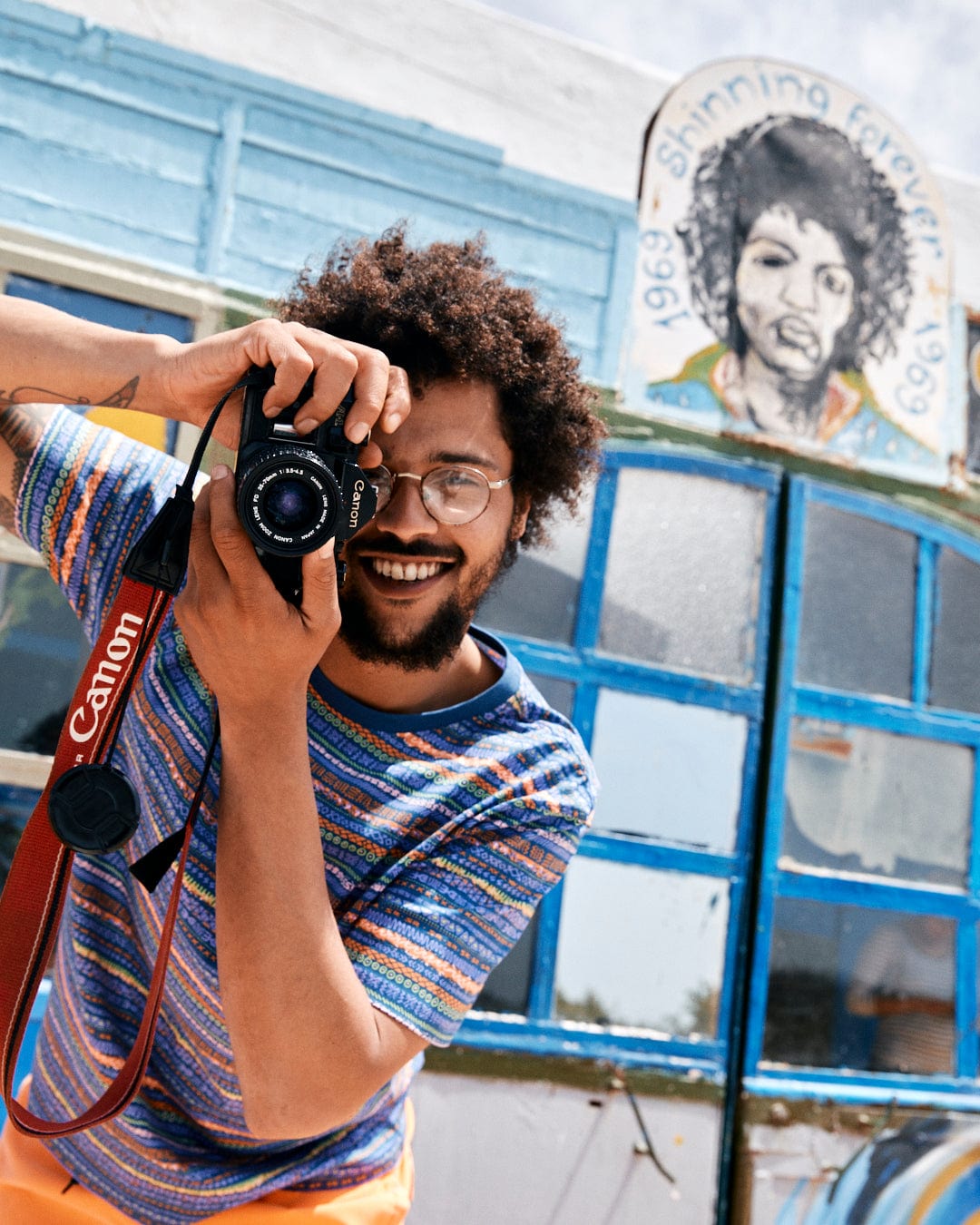 A smiling man with curly hair taking a photo with a Canon camera, standing in front of a colorful Aztec print mural of a face wearing a Saltrock Marks - Mens Short Sleeve T-Shirt in Purple.