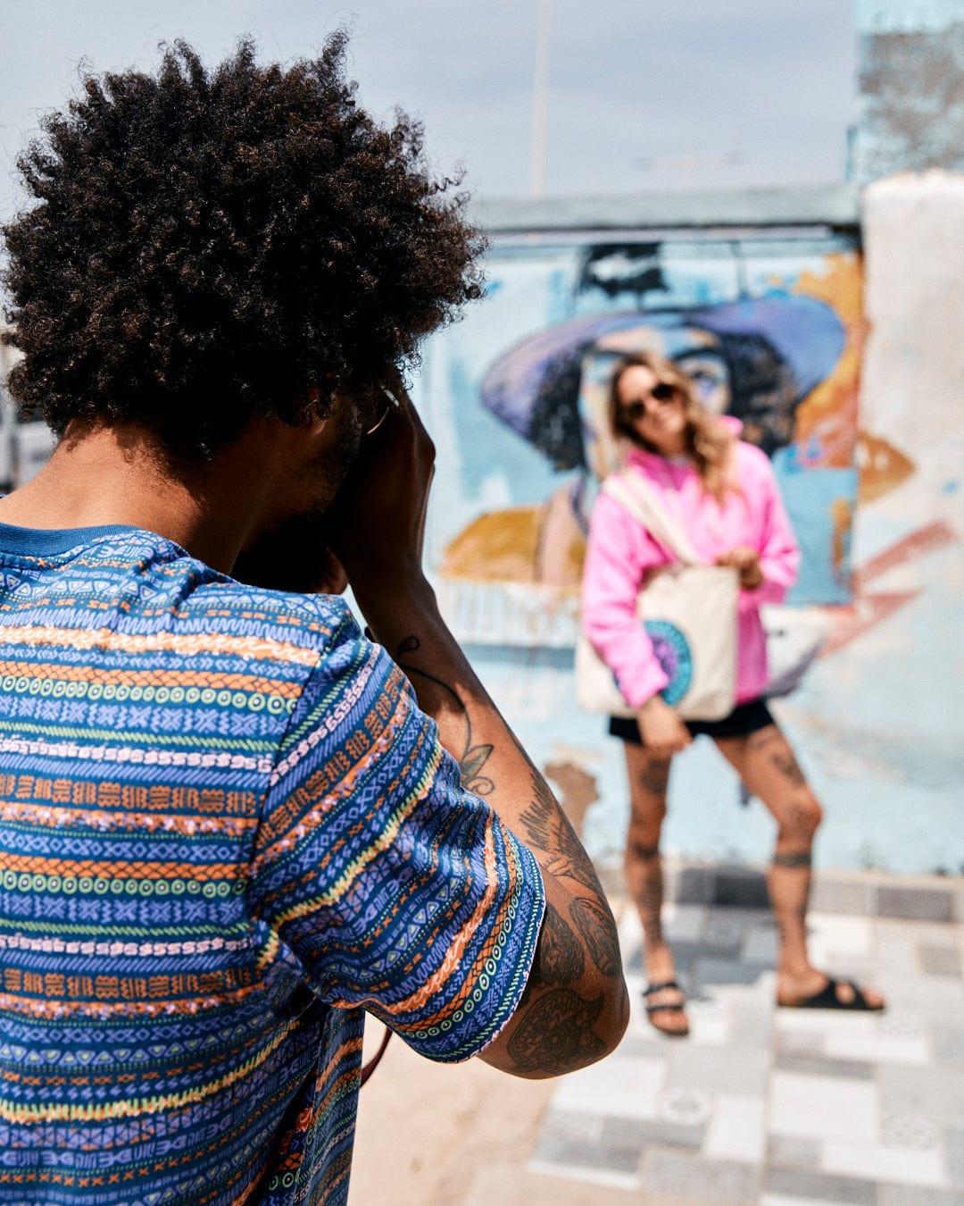 A man takes a photo of a woman posing in front of a vibrant street mural; she is smiling and holding a hat with Aztec print, and wearing the Saltrock Marks Men's Short Sleeve T-Shirt in Purple.