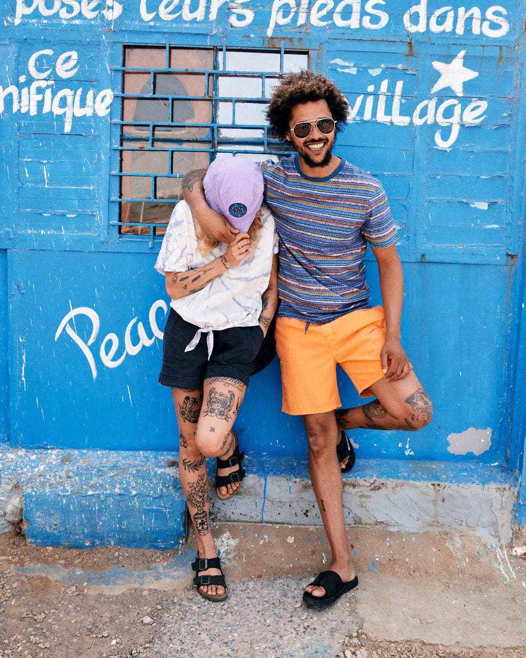 A couple with tattoos poses in front of a blue wooden wall with Aztec print graffiti, laughing, with the man lifting one leg and wearing sunglasses while wearing Marks - Mens Short Sleeve T-Shirt in Purple by Saltrock.