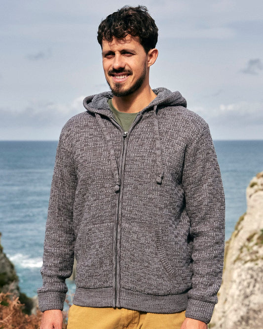 A man modeling the Saltrock branding, wearing a Markus - Mens Borg Lined Knit Hoodie - Dark Grey for warmth, stands next to the ocean.