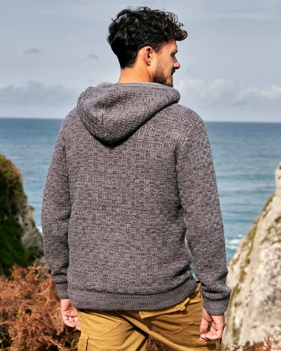 A man wearing a Saltrock Markus - Mens Borg Lined Knit Hoodie - Grey, embracing winter days as he gazes at the ocean.