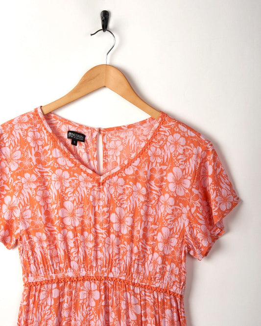 Marla Hibiscus dress in pink/orange by Saltrock hanging on a wooden hanger against a white wall.
