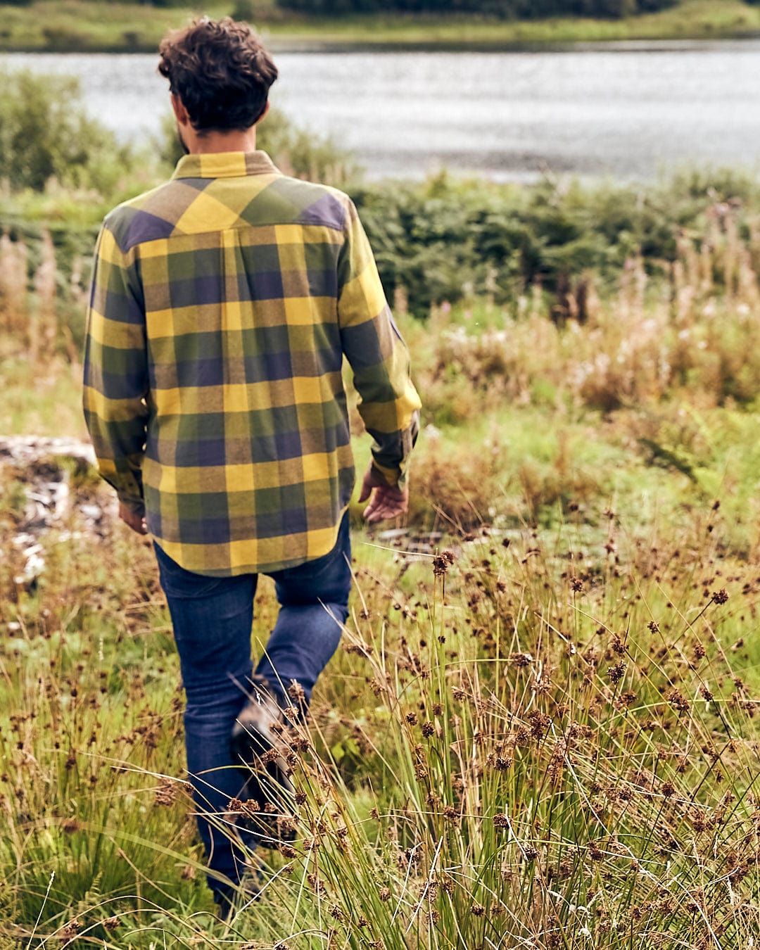 A man in a Saltrock Mattie - Mens Long Sleeve Shirt - Yellow walks through a field near a lake.