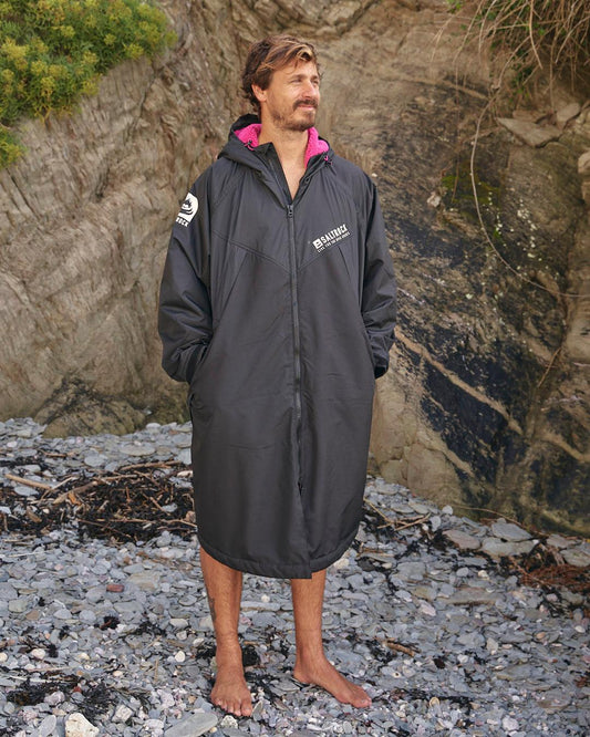 A man in a Saltrock Four Seasons - Waterproof Changing Robe - Black/Pink standing on a rocky beach.