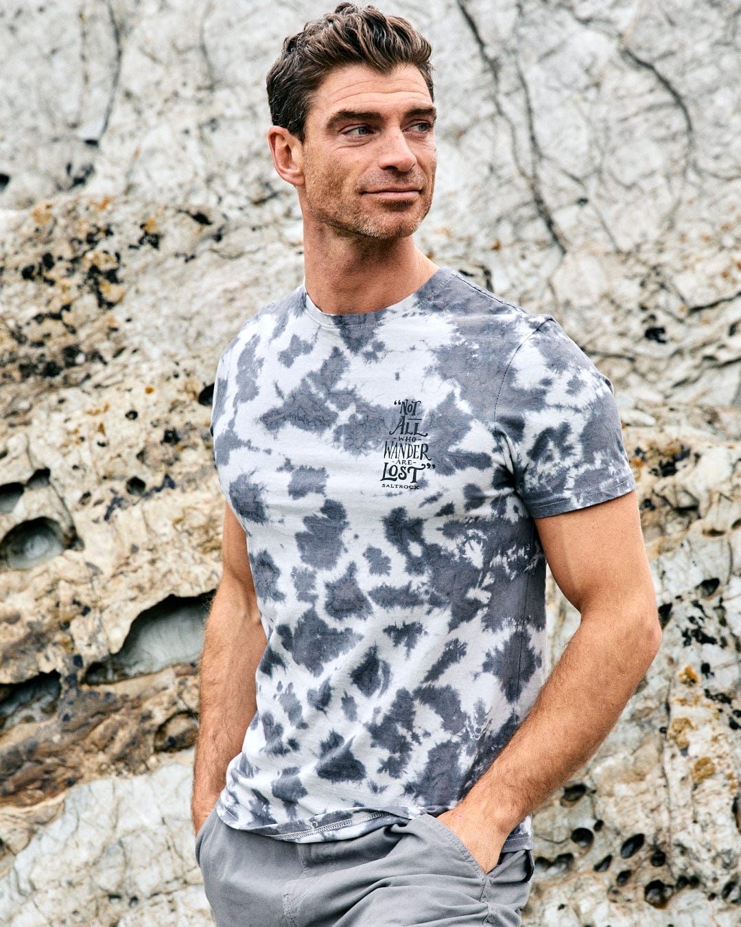 A man stands before rocky terrain, wearing the Saltrock Lost Ships Men's Tie Dye T-Shirt in grey and white, featuring "Not All Who Wander Are Lost" text on the chest.