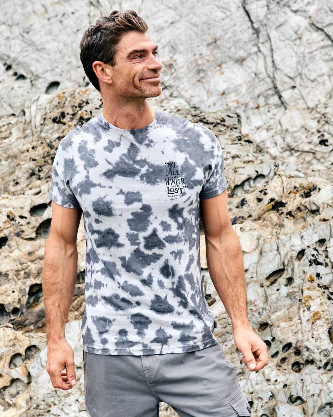 A man in the Saltrock Lost Ships Men's Tie Dye T-Shirt in grey stands outdoors before rocky scenery, glancing to the side and smiling.