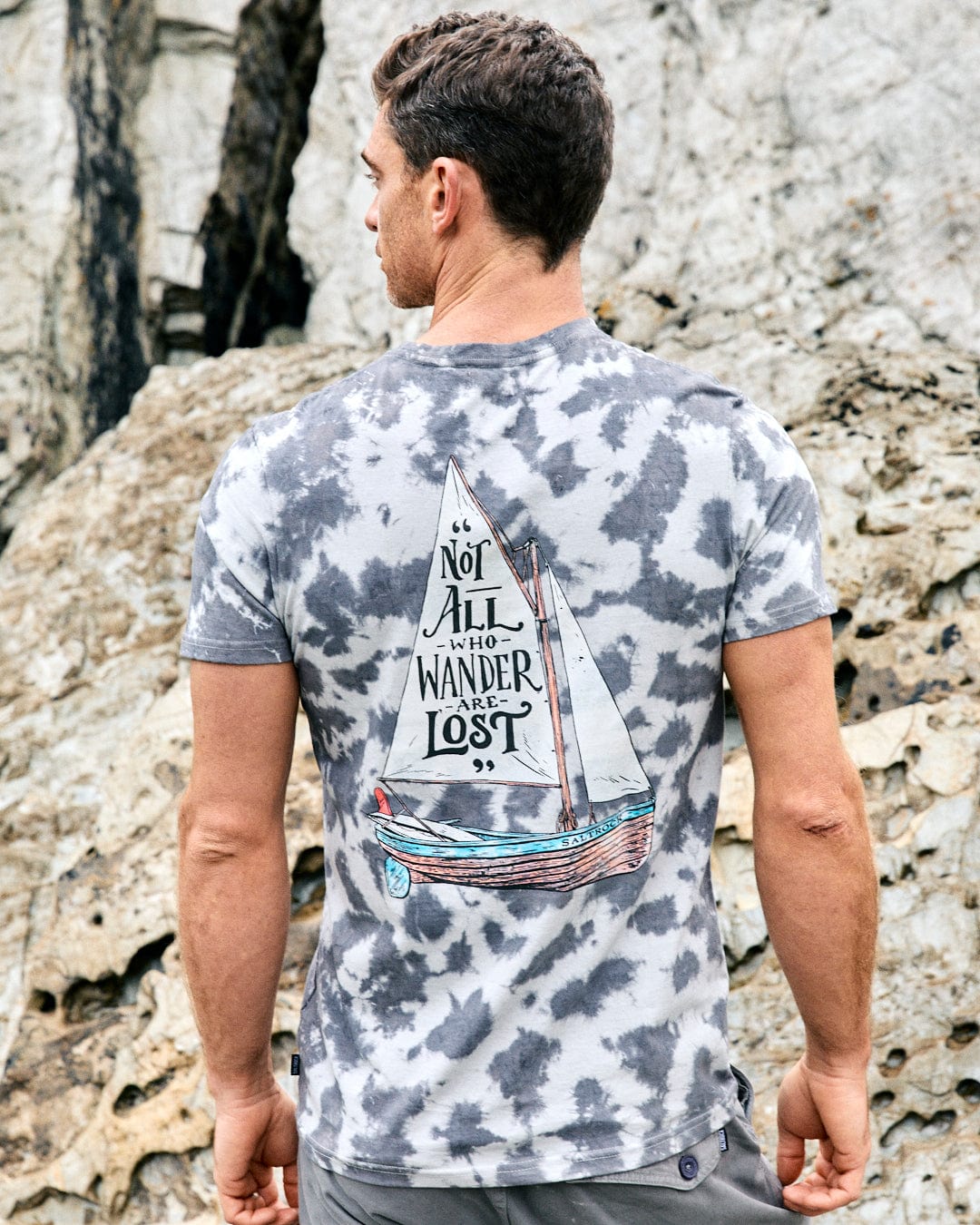 A man stands with his back to the camera, wearing the Saltrock Lost Ships men's grey tie-dye t-shirt with a sailboat graphic and "Not all who wander are lost" text. Rocky background.