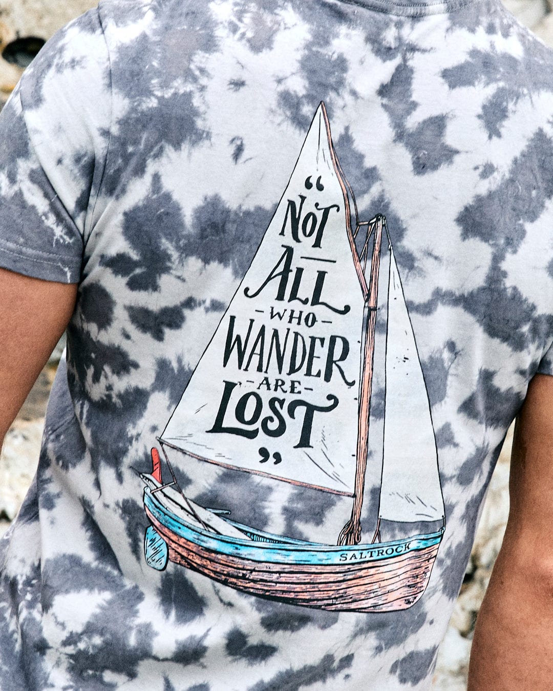 A person wears the Saltrock Lost Ships men's grey tie-dye T-shirt, featuring a sailboat graphic and “Not all who wander are lost” on the back.