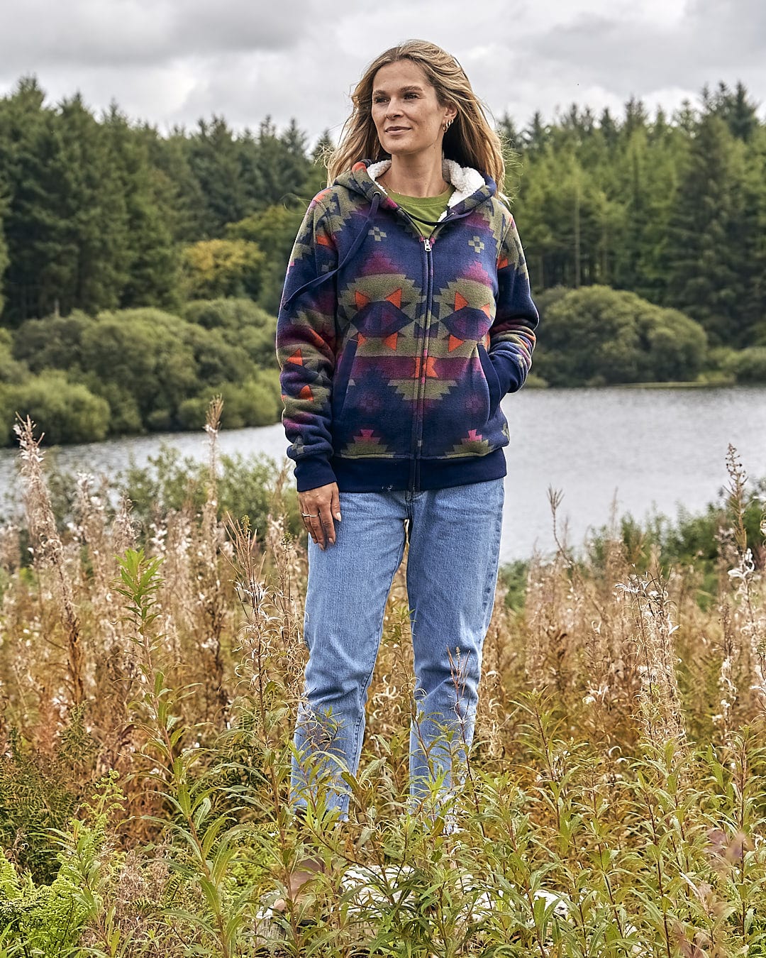 A woman standing in a field next to a lake wearing a Saltrock Merida - Womens Lined Aztec Zip Fleece - Dark Blue hoodie.