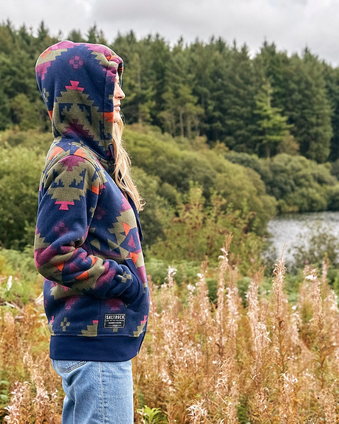 A woman wearing a Saltrock hoodie standing in a field near a lake.