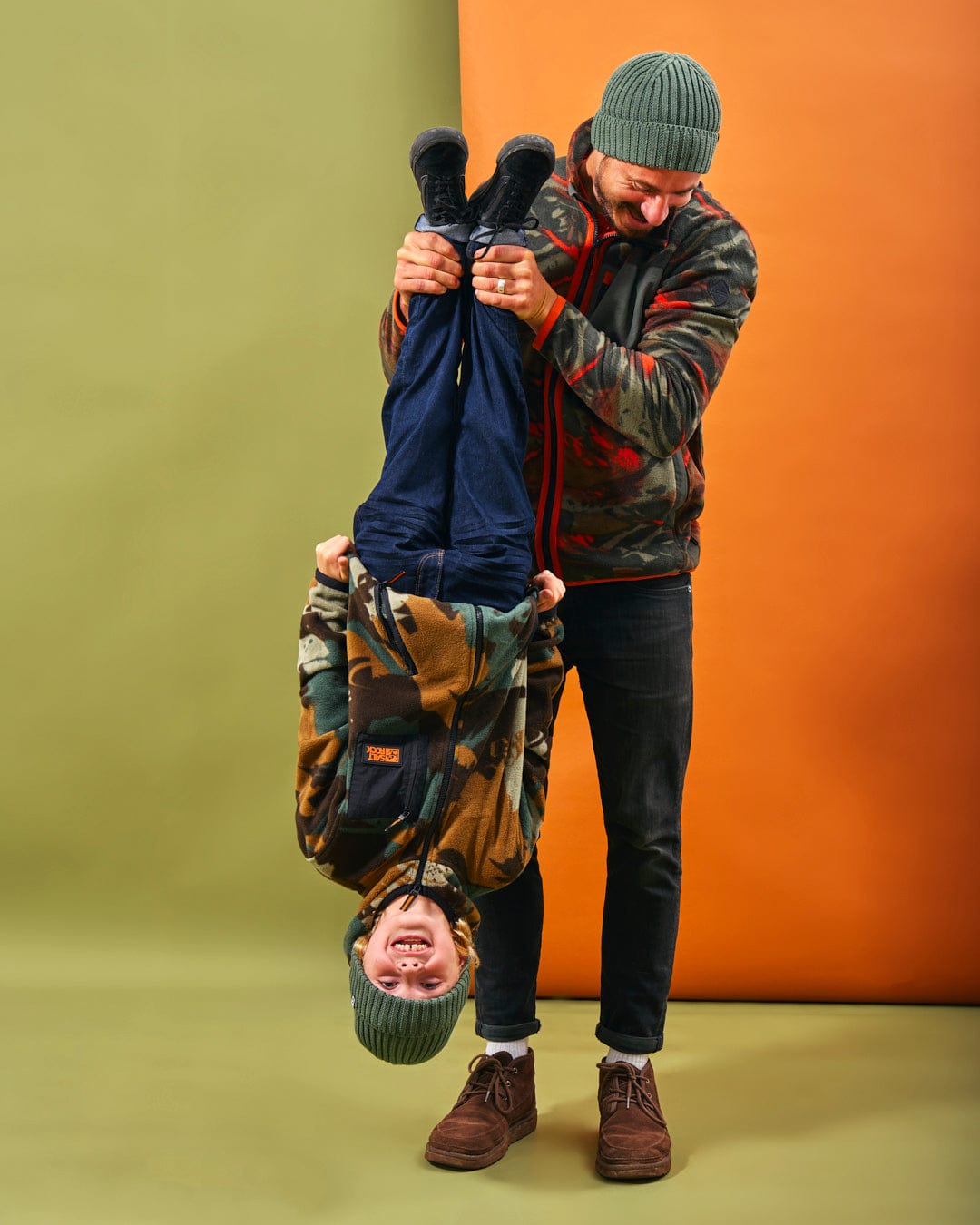 A man is holding a boy upside down in front of an orange background while they both wear Saltrock's Communicado - Kids Camo Zip Fleece - Dark Green jackets with front zip.