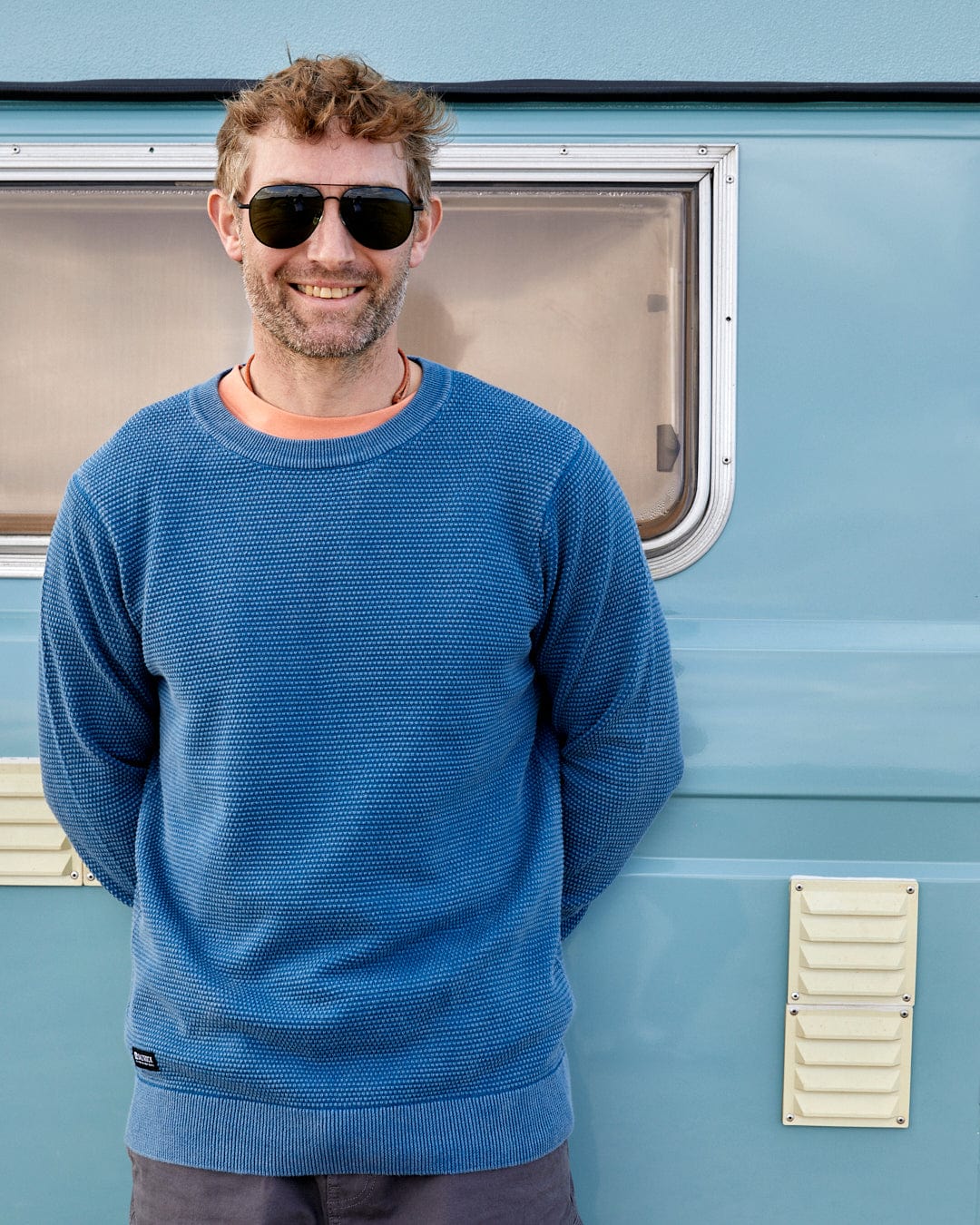 A man in a Saltrock Moss - Mens Washed Knitted Crew - Blue cotton sweatshirt standing next to a blue camper van.