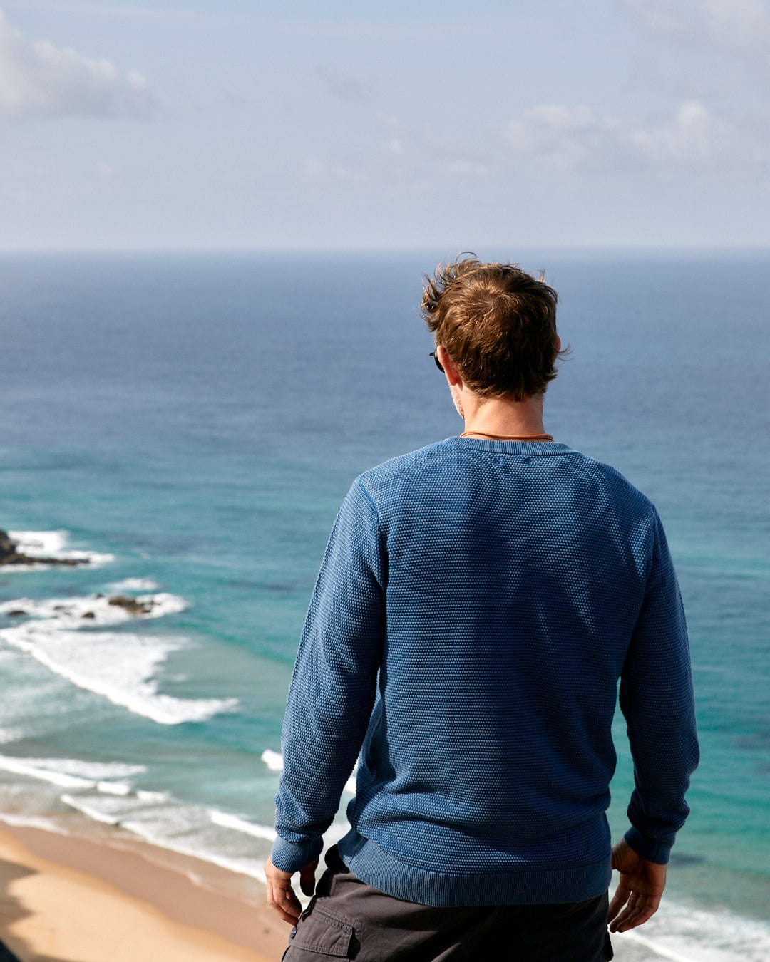 A man standing on a Moss - Mens Washed Knitted Crew - Blue cliff overlooking the blue ocean. (Saltrock)