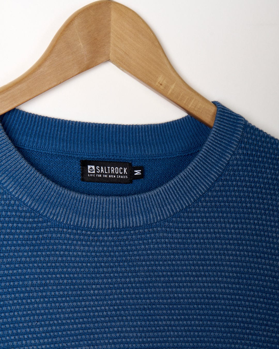 A Saltrock, Moss - Mens Washed Knitted Crew - Blue sweater hangs on a wooden hanger.