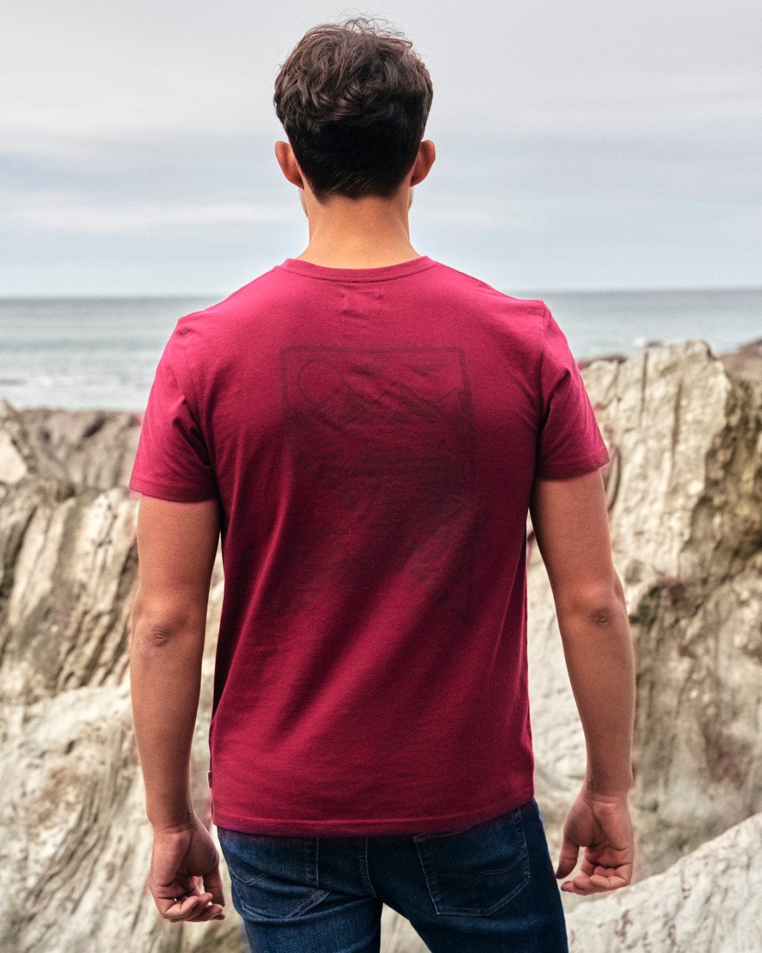 A man wearing a maroon Saltrock T-Shirt is looking at the ocean.