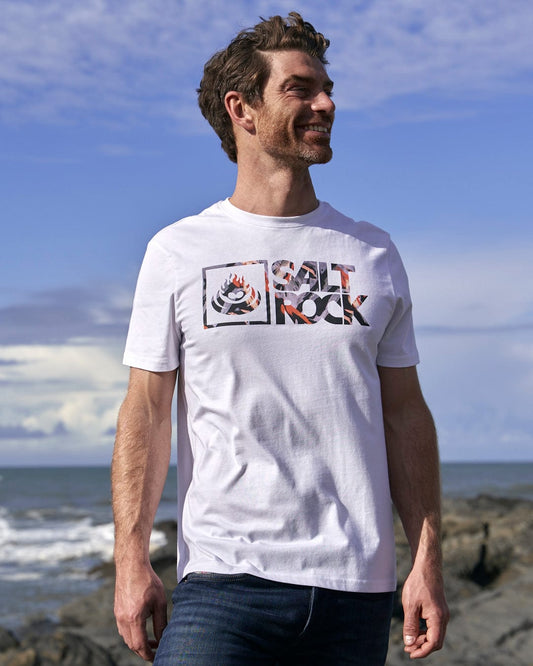 A man wearing a white t-shirt standing on rocks, showcasing the Saltrock Mountain Logo - Mens Short Sleeve T-Shirt - White branding.