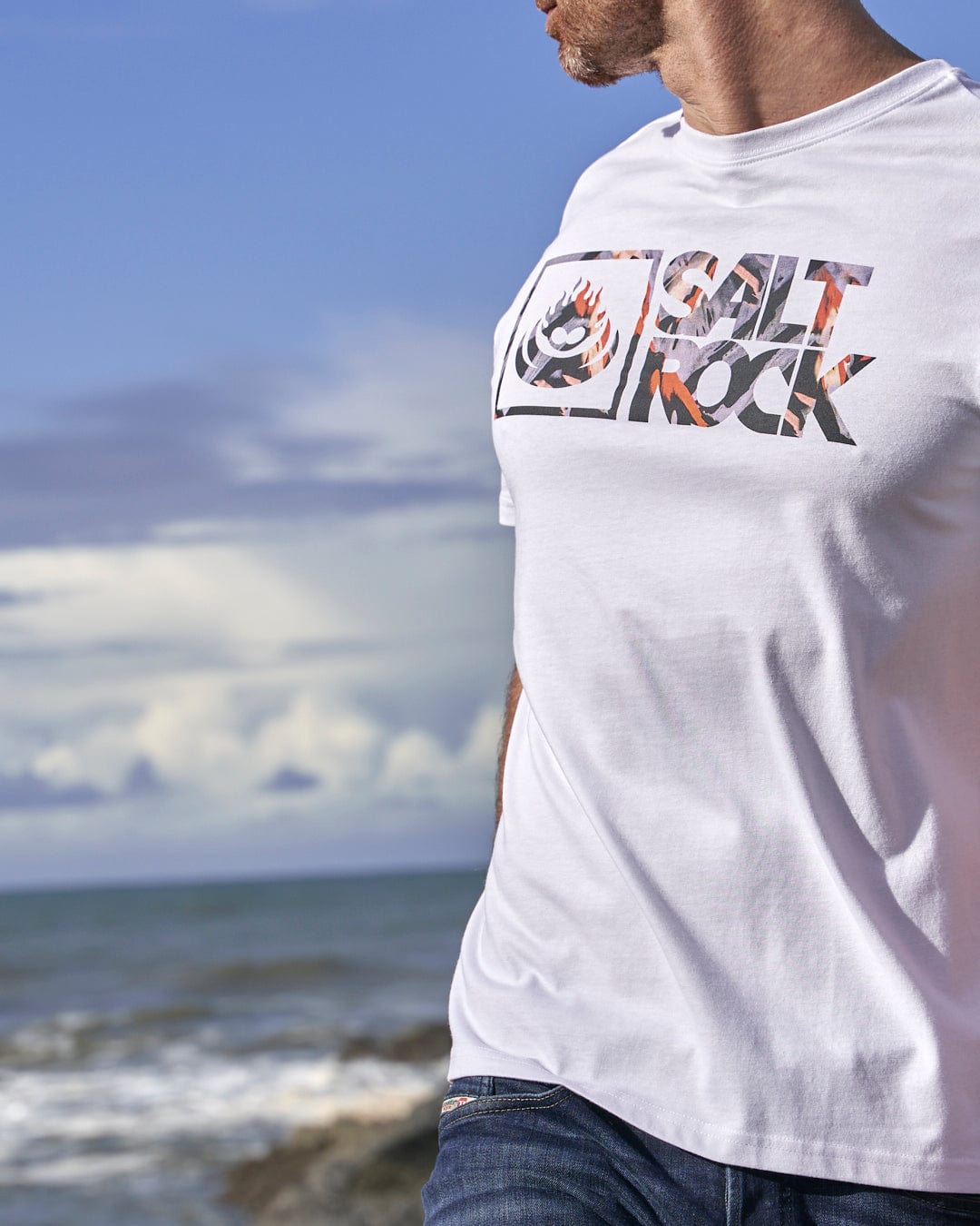 A man is standing on the beach wearing a white Mountain Logo - Mens Short Sleeve T-Shirt featuring the Saltrock branding.