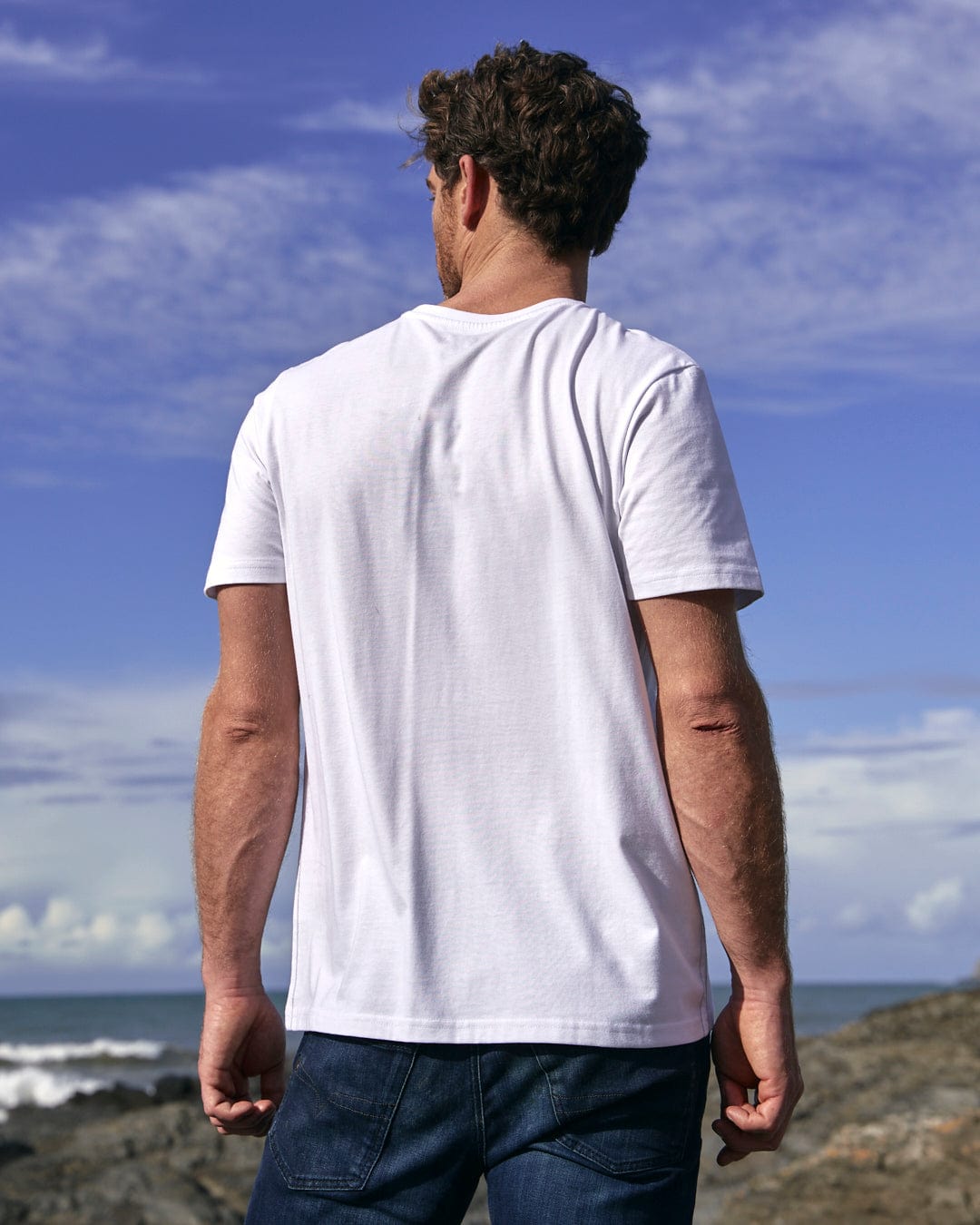 A man wearing a white Mountain Logo - Mens Short Sleeve T-Shirt - White featuring the Saltrock branding, standing on a rocky beach.