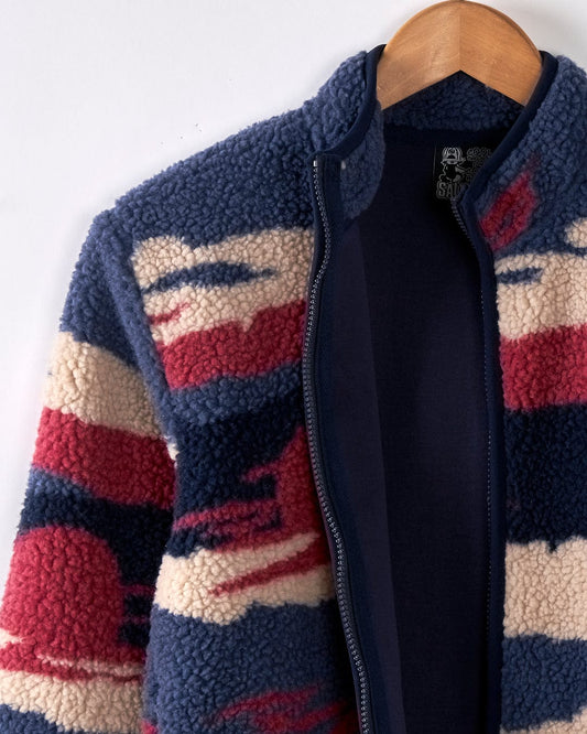 The "Mountain Trail - Kids Recycled Borg Zip Fleece - Blue" by Saltrock hangs on a wooden hanger. It showcases a textured pattern in navy, red, and cream colors, featuring a warm recycled cosy sherpa lining. The sherpa-lined collar offers comfort while maintaining style and sustainability.