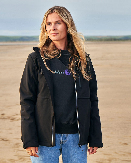 A woman wearing a Saltrock Solae 2 - Womens Softshell Jacket - Black standing on a beach.