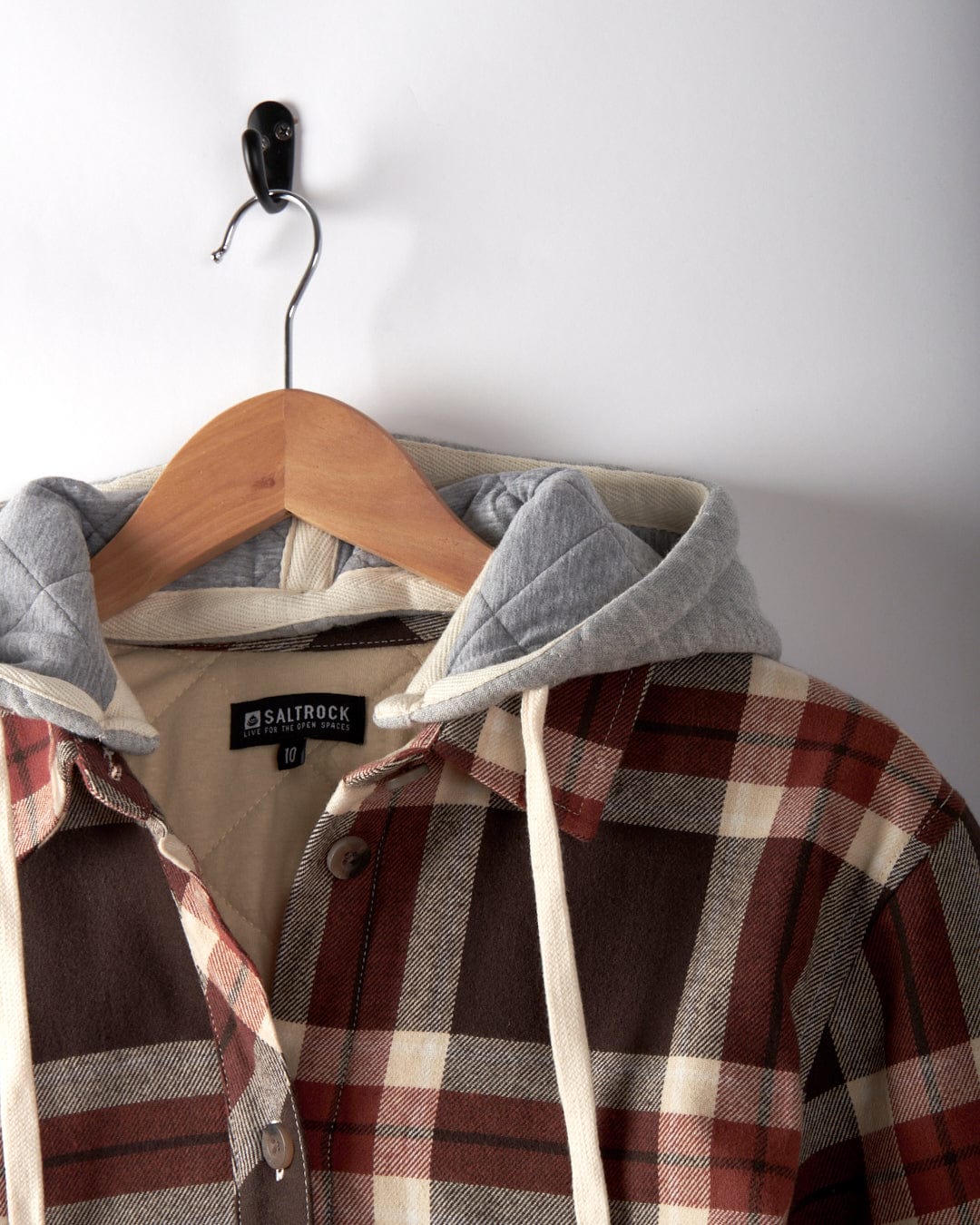 The Saltrock Myla Womens Hooded Shacket in brown features a check plaid pattern, oversized fit, and a gray detachable hood, shown hanging on a wooden hanger from a metal wall hook.