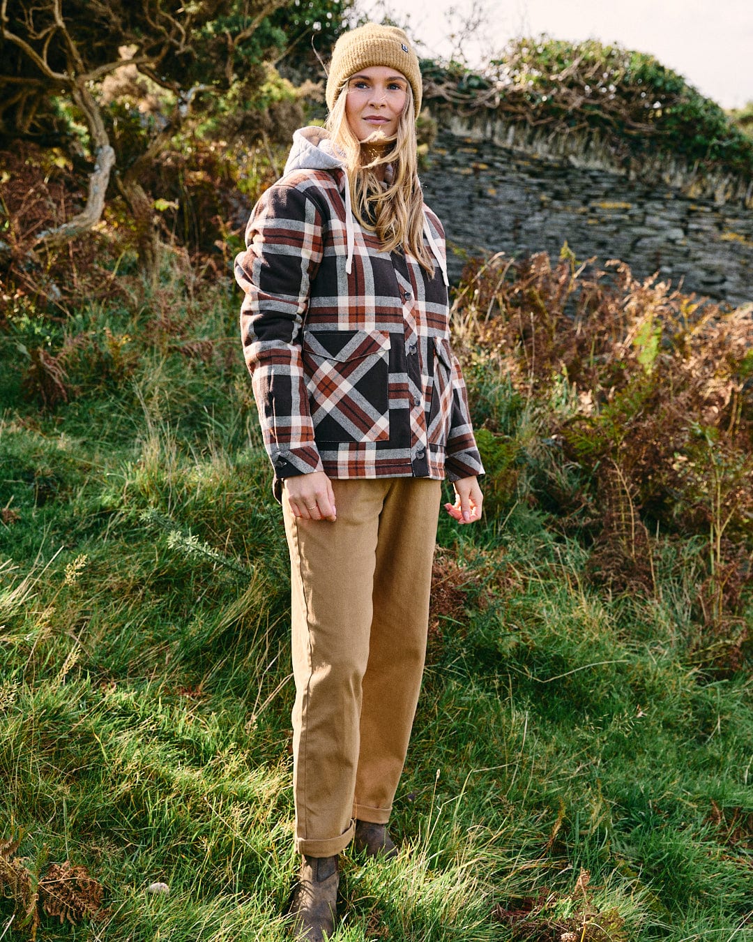 A person wears the Saltrock Myla Women's Hooded Shacket in brown, paired with tan pants and a yellow knit beanie, standing outdoors on grass with a stone wall and greenery in the background.
