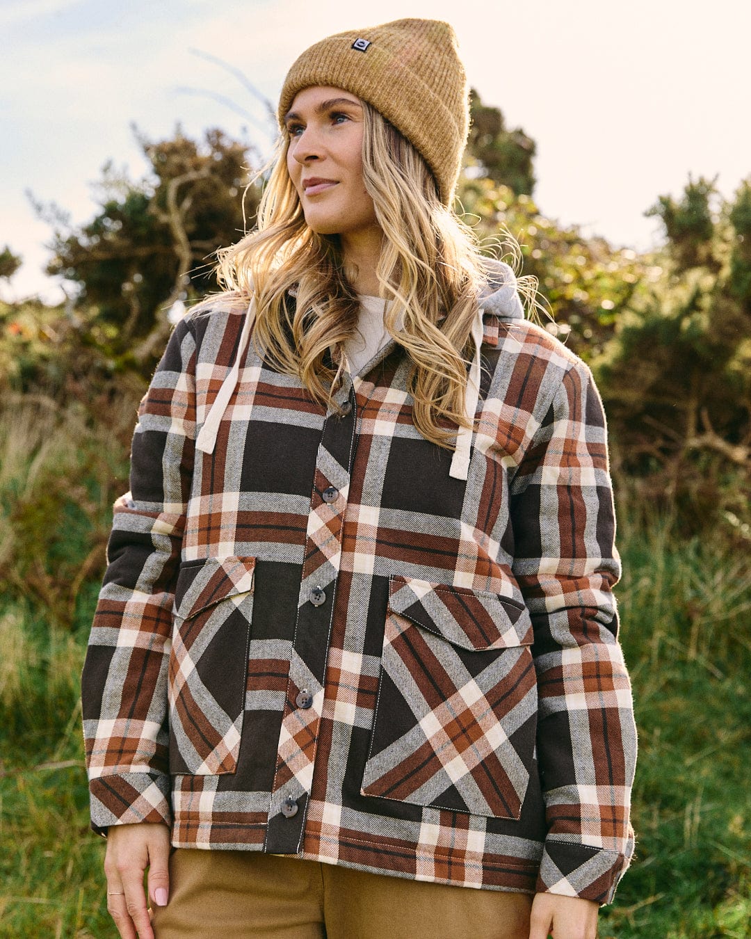 A woman stands outdoors in sunlight and greenery, wearing the Saltrock Myla Women's Hooded Shacket in brown with a tan beanie.