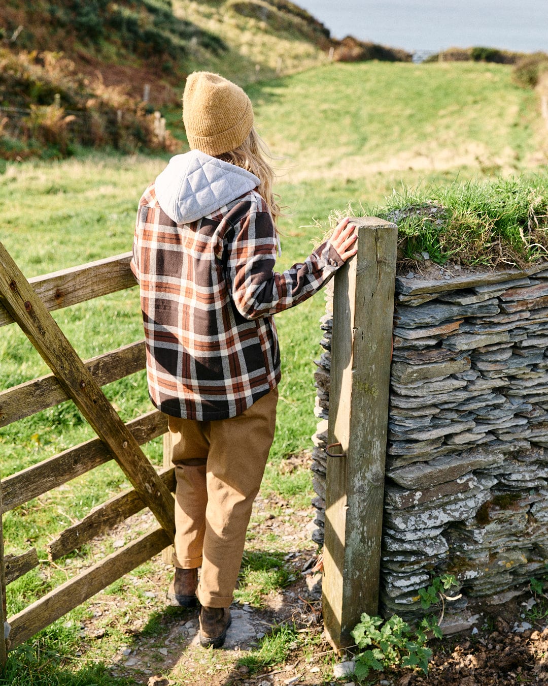 A person wearing the Saltrock Myla Women's Hooded Shacket in brown and a tan beanie stands by a wooden gate, gazing over grassy hills in the distance.