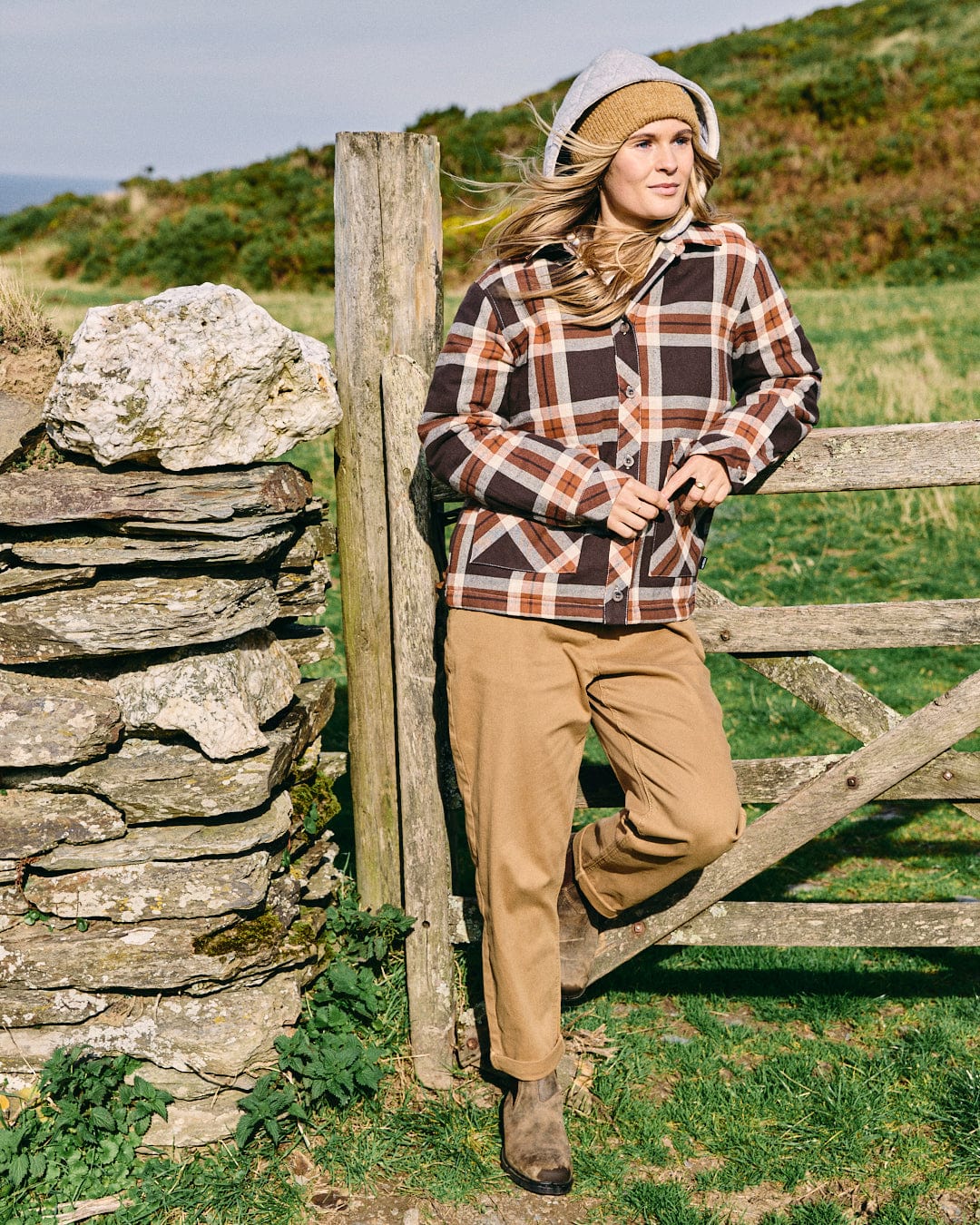 Wearing the Saltrock Myla Women's Hooded Shacket in brown, a person leans against a wooden gate by a stone wall, dressed in tan pants and boots in a rural outdoor setting.