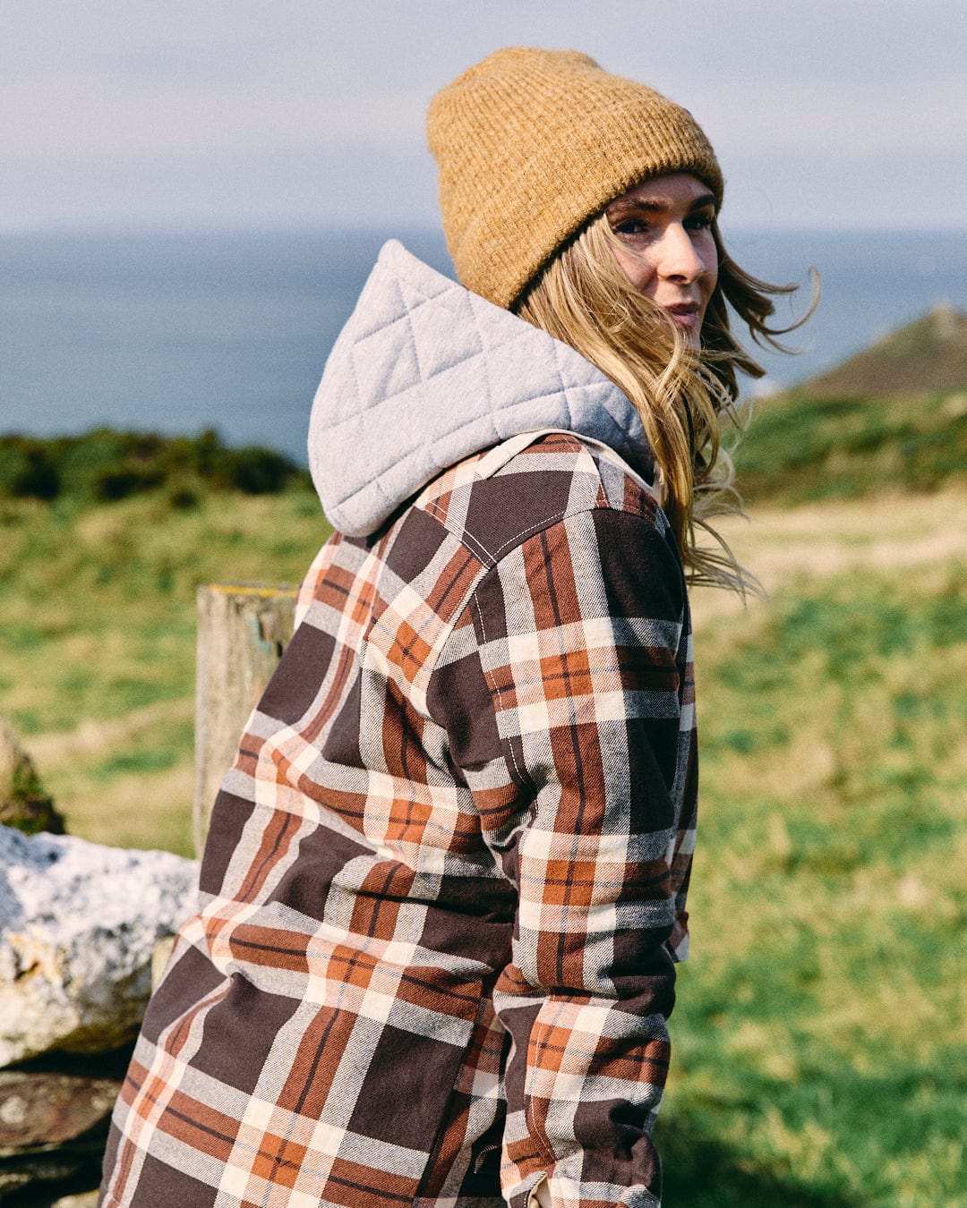 A woman wears the Saltrock Myla Women's Hooded Shacket in brown and a tan knit beanie, standing outdoors on grass with hills and water in the background.