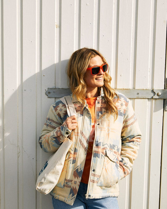 A person wearing a Saltrock Mylene - Aztec Shacket - Cream and red sunglasses stands against a light-colored wooden wall, holding a tote bag and smiling.