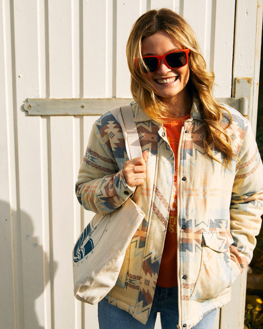 A person wearing a Saltrock Mylene - Aztec Shacket - Cream with red sunglasses smiles while standing in front of a white wooden door, holding a tote bag.