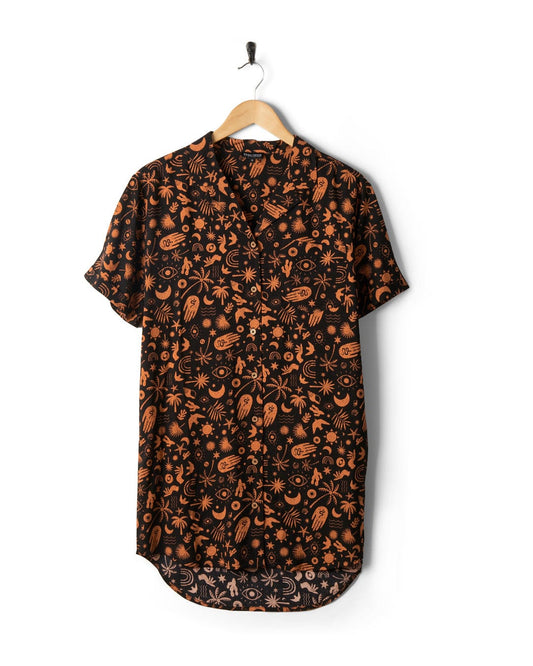 Saltrock Mystic Tattoo - Womens Shirt Dress - Black features an orange mystical tattoo style print with various symbols, such as stars, moons, and hands on a black short-sleeve shirt. Made of Soft Spun Viscose, it's both lightweight and hangs effortlessly on a wooden hanger against a white background.