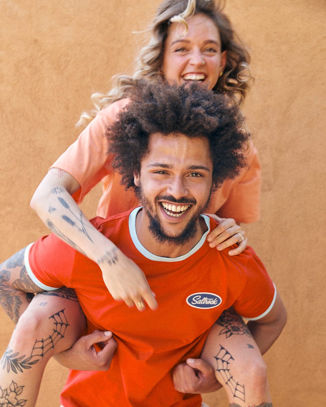 A joyful woman piggybacking on a cheerful man, both with tattoos and wearing red Striker Ringer - Mens Short Sleeve T-Shirts with Saltrock embroidered branding, smiling against a tan background.