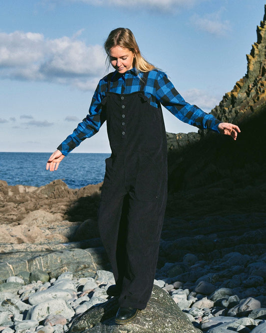 A stylish woman wearing Nancy - Womens Cord Dungaree - Dark Blue overalls confidently stands on rocks by the ocean.