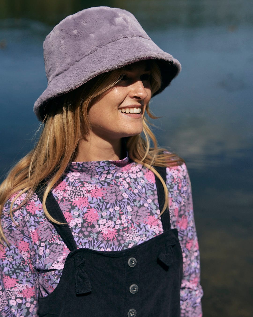 A stylish woman in overalls and a hat is standing by a body of water, rocking her Saltrock - Brooklyn Womens Long Sleeve T-Shirt - Purple.