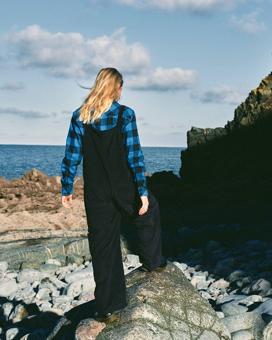 A fashion-conscious woman in stylish Saltrock Nancy - Womens Cord Dungaree - Dark Blue, standing on rocks near the ocean.