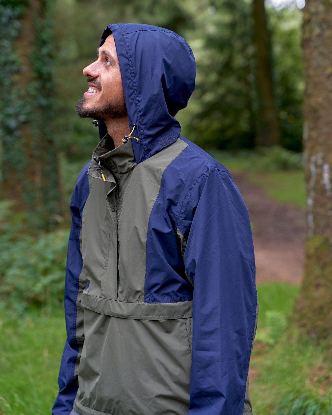 A man wearing a Saltrock Nevis - Mens Waterproof Mac - Blue/Green jacket standing in a wooded area.