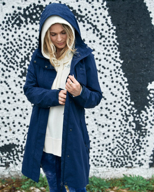 A woman in a Saltrock North West blue hooded waterproof jacket standing in front of a black and white abstract mural, buttoning her coat.