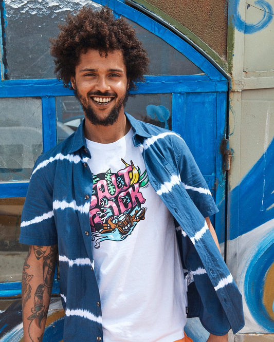 A cheerful man with curly hair, wearing a Saltrock Ocean - Mens Tie Dye Shirt - Blue and an open blue flannel shirt, stands in front of a colorful, graffiti-covered door.