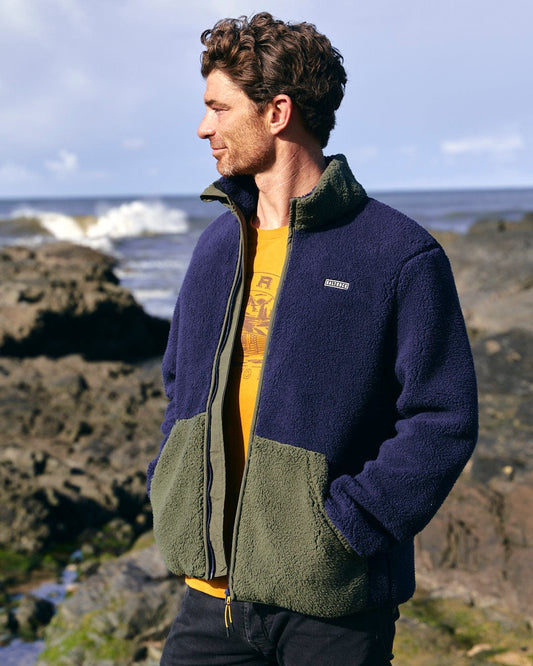 A man is standing on rocks near the ocean wearing a Saltrock Odin -  Mens Zip Thru Fleece - Dark Blue jacket.