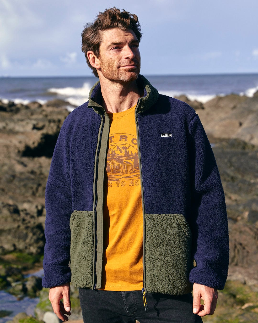 A man is standing on the beach wearing a colourblocked blue and yellow jacket, specifically a Saltrock Odin - Mens Zip Thru Fleece.