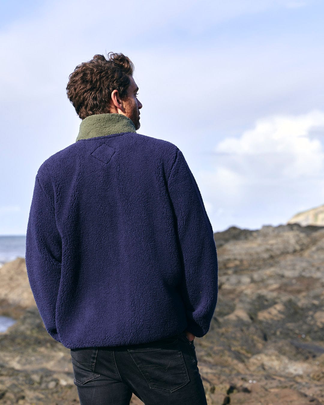 A man wearing a blue sweater, Saltrock Odin - Mens Zip Thru Fleece, standing on a rocky beach.