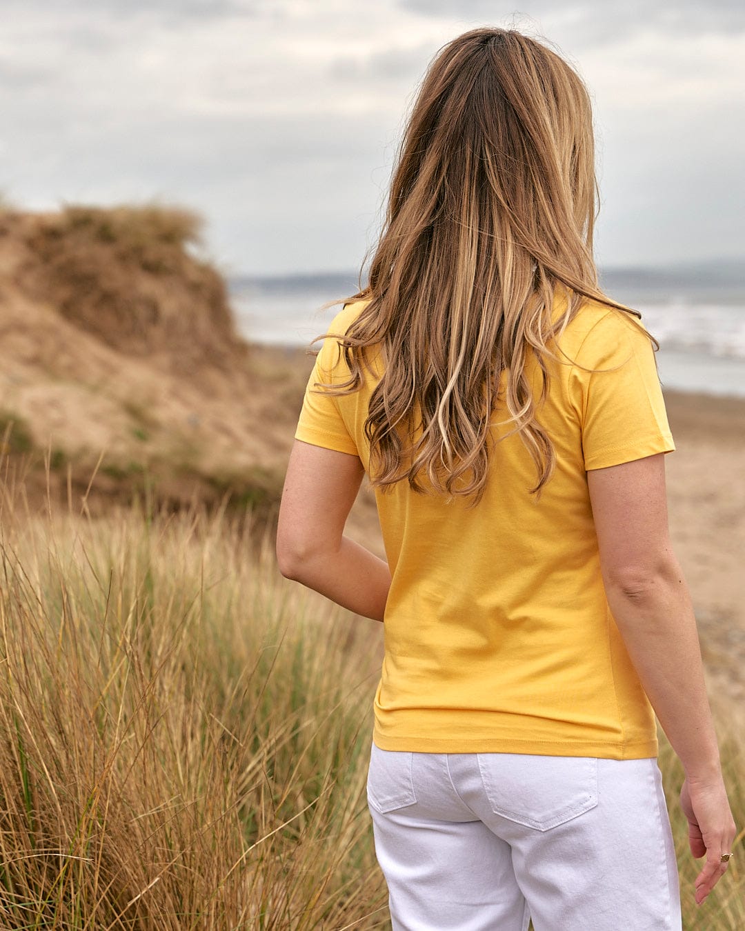 A woman wearing a Saltrock On The Road Cornwall - Womens Short Sleeve T-Shirt - Yellow standing on the beach.
