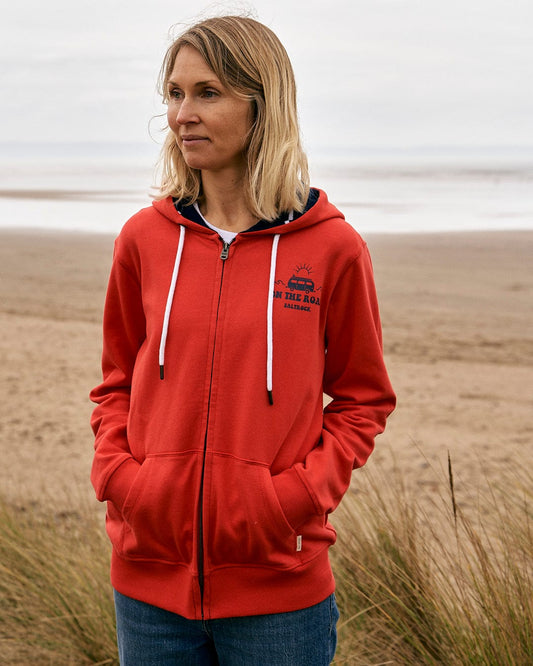 A woman wearing a Saltrock On The Road Cornwall - Womens Zip Hoodie - Red standing on a beach.