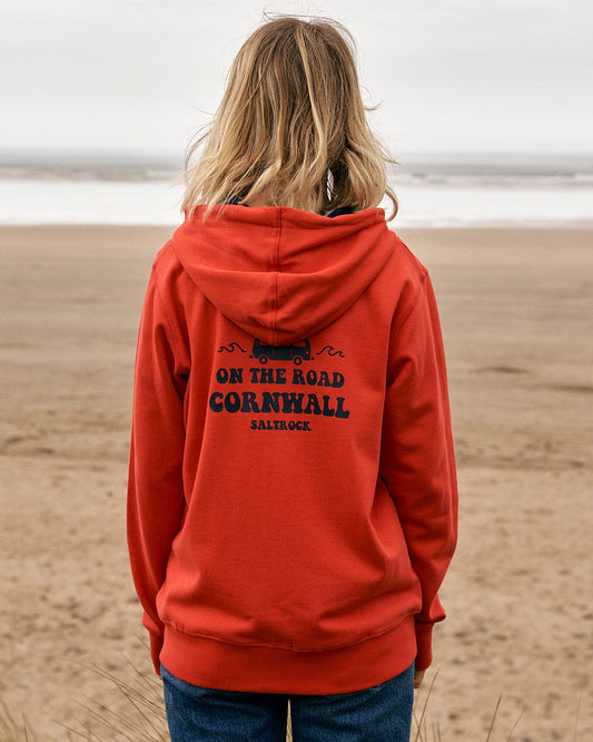A woman is standing on the beach wearing a Saltrock On The Road Cornwall - Womens Zip Hoodie - Red.