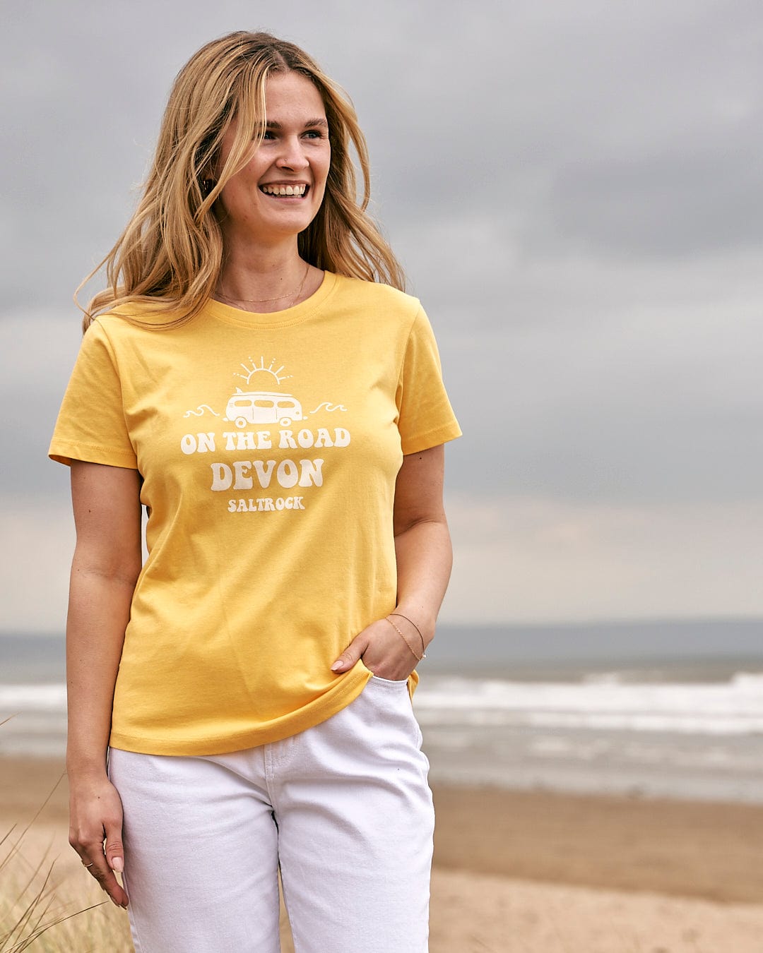 A woman wearing a Saltrock On The Road Devon - Womens Short Sleeve T-Shirt - Yellow standing on the beach.