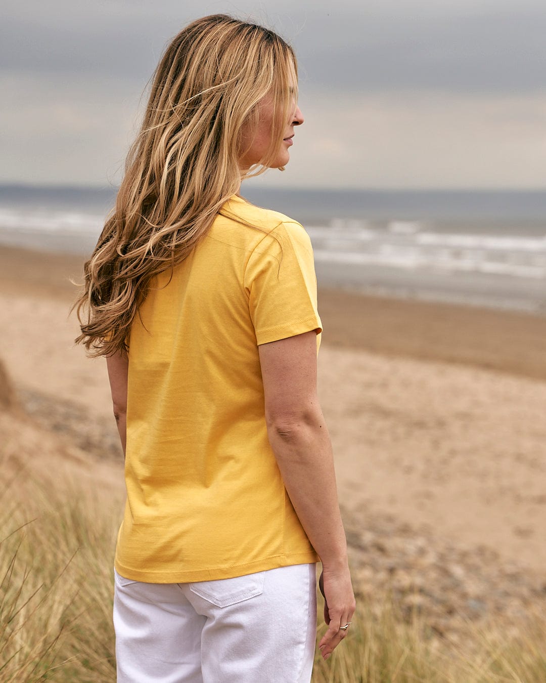 A woman wearing the Saltrock On The Road Devon - Womens Short Sleeve T-Shirt - Yellow is standing on the beach.